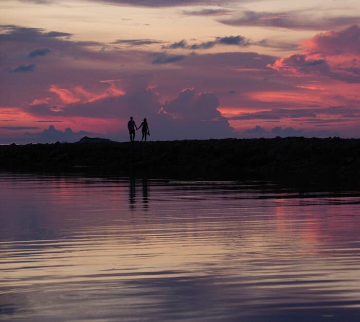 Silhouette of a couple holding hands by the lakeshore at a stunning sunset, reflecting vibrant colors.